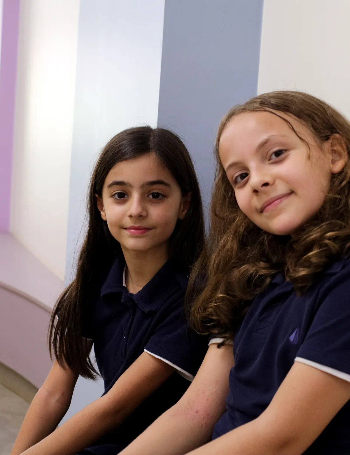 Duas alunas sentadas lado a lado em um banco do corredor da escola