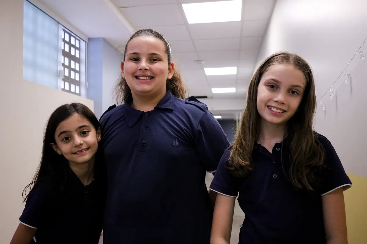 Três alunas abraçadas sorrindo no corredor da escola