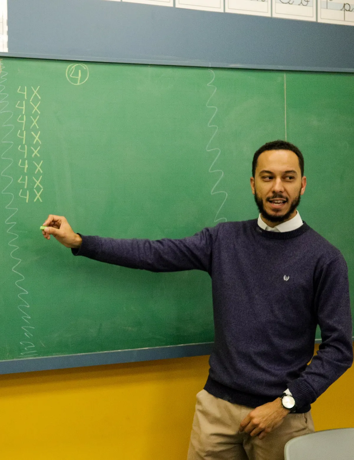 Professor explicando na lousa verde, com alunos de costas