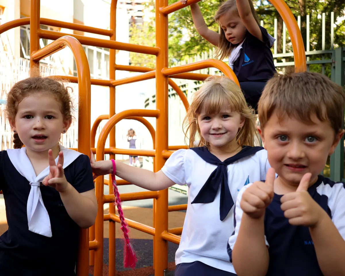 Três crianças da Educação Infantil fazendo joinha no trepa-trepa laranja do parque